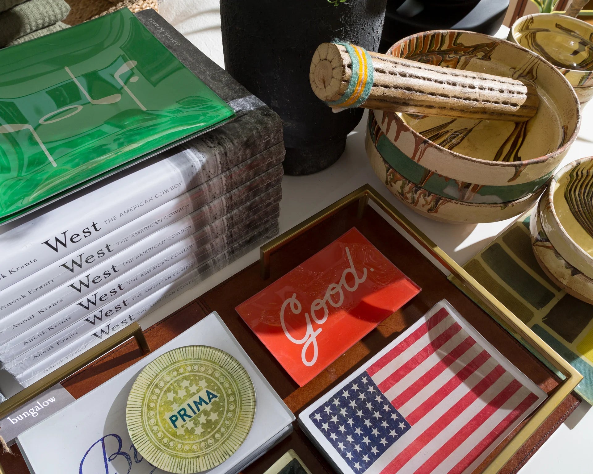 A table with books, decorative trays, a small American flag, ceramic bowls, and a pestle. Items include a stack of "West" books and a tray with the word "Good.