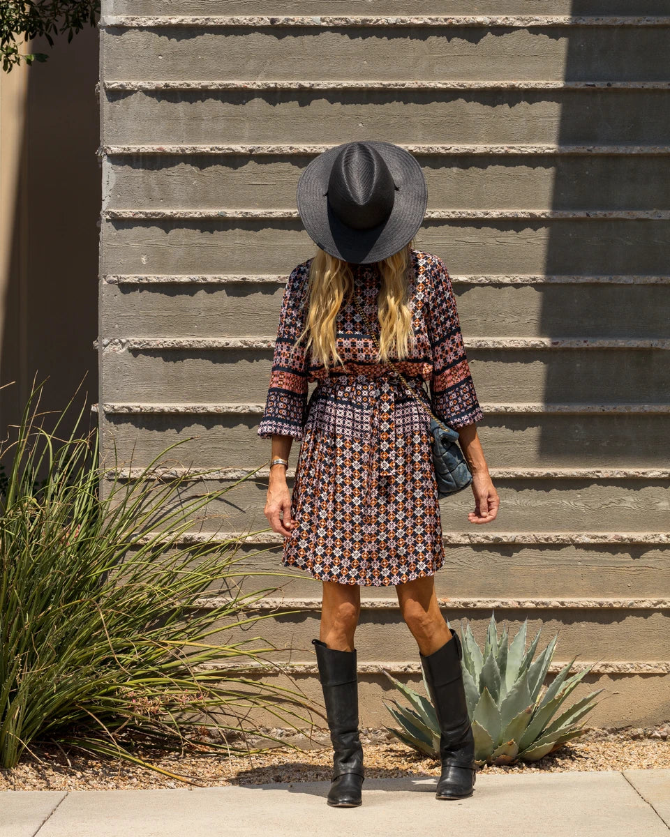 A woman with long blonde hair wearing a wide-brimmed black hat, patterned dress, and knee-high black boots stands in front of a textured concrete wall. Her face is obscured by the hat. There are green plants near her feet.