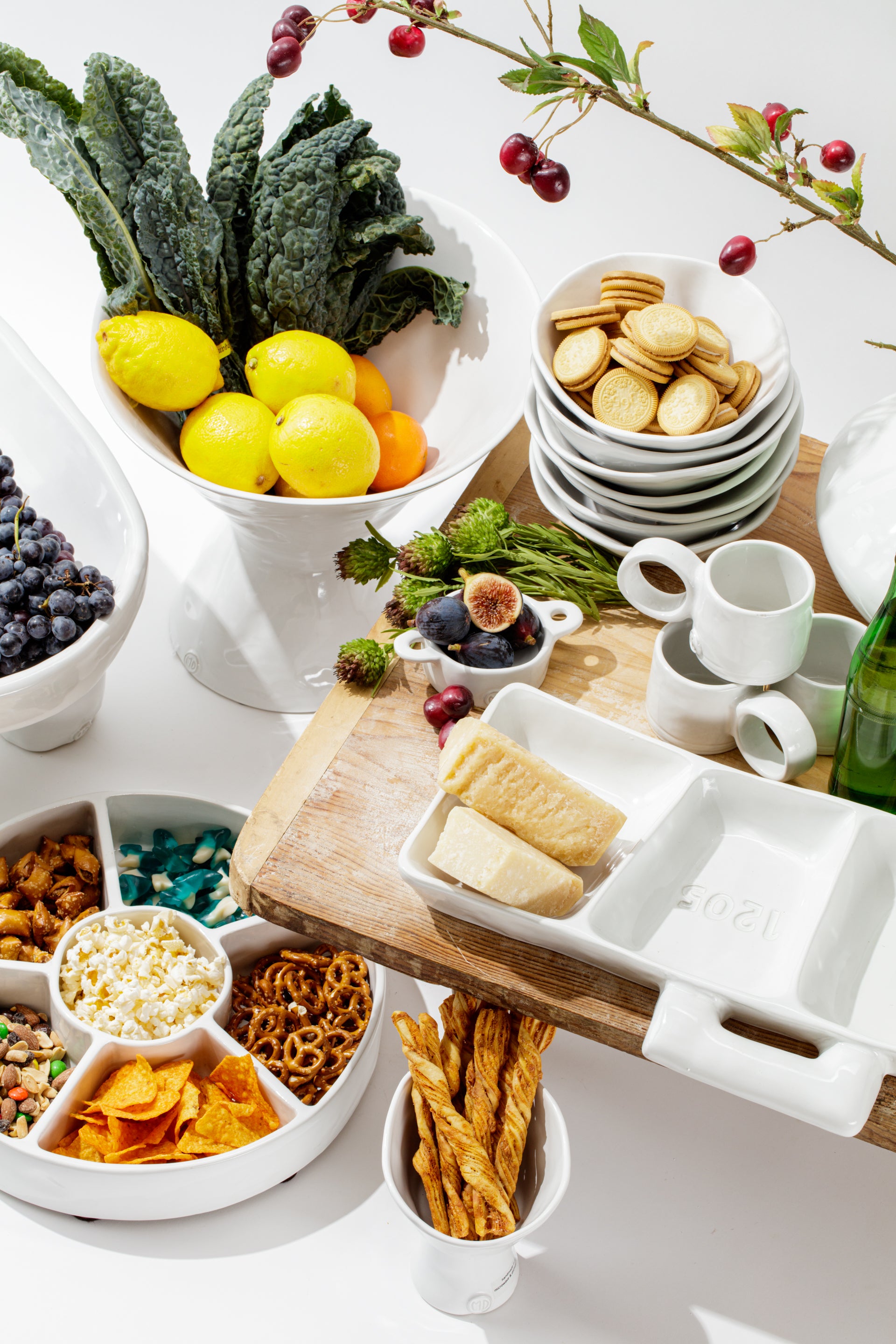 A vibrant assortment of food displayed artistically. Includes a bowl of fresh kale, lemons, a tray with cookies, a dish with assorted snacks like popcorn and pretzels, and other fruits and herbs, all on a white table.