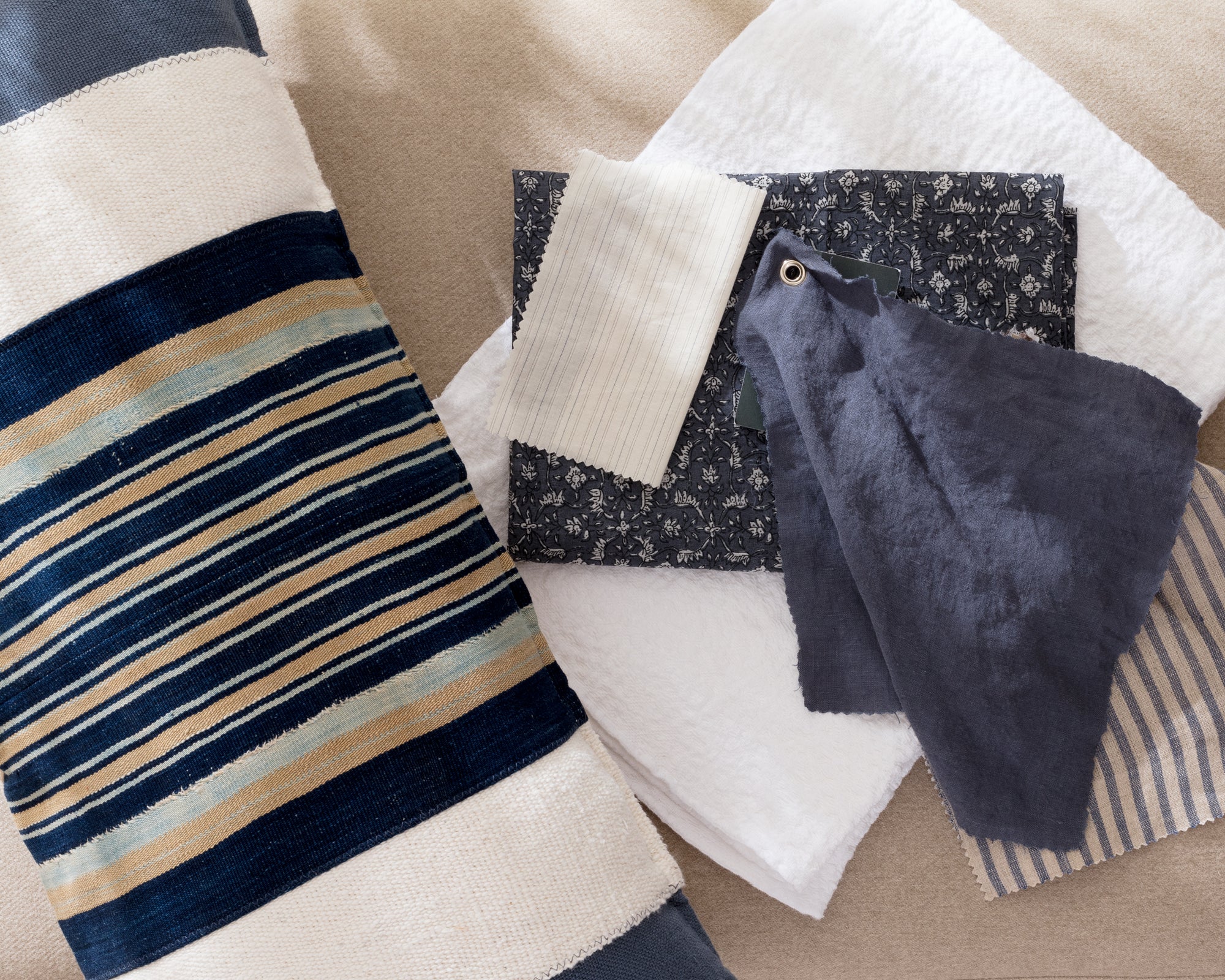 A striped pillow with blue, beige, and white tones lies next to various fabric swatches in navy, cream, and patterned prints, all arranged on a neutral-colored surface.