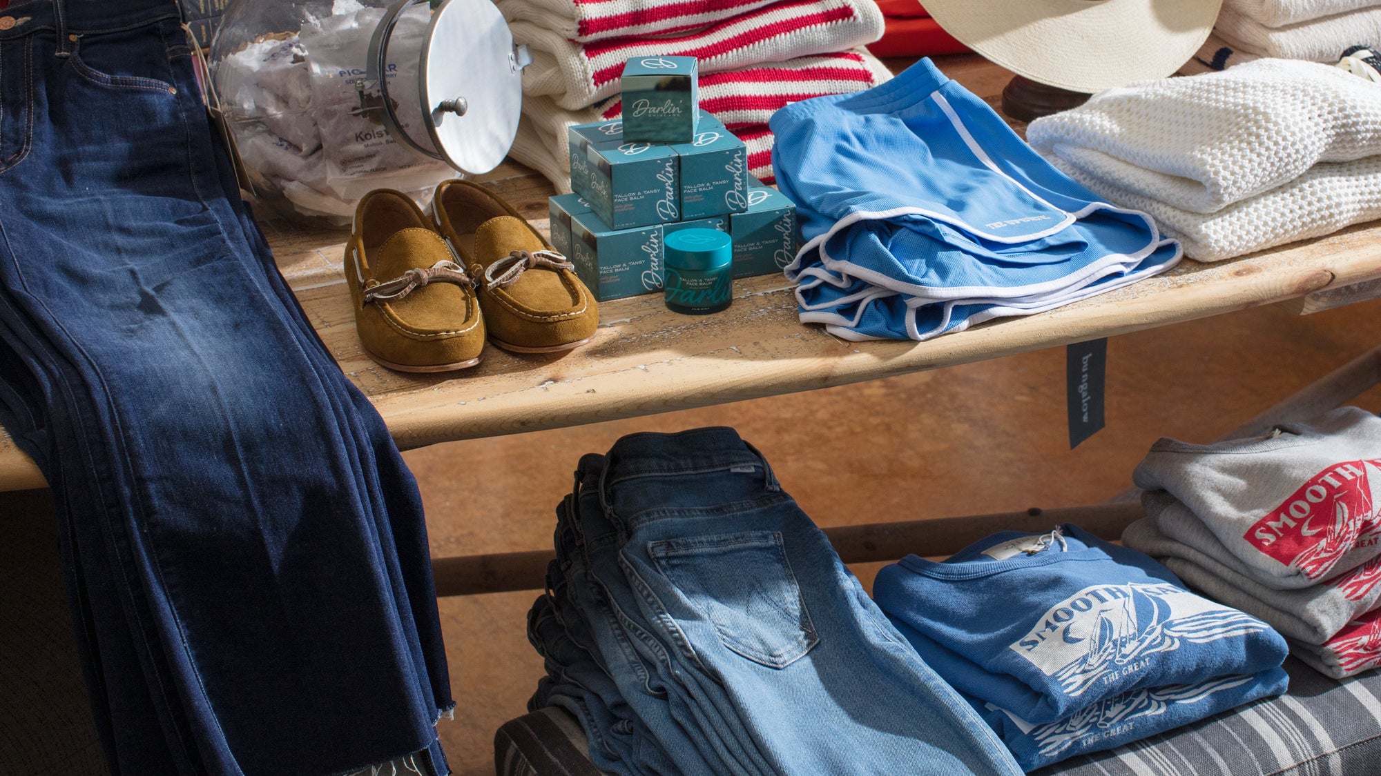 A wooden table displays folded jeans, a striped shirt, white sweater, blue boxer briefs, brown loafers, grooming products, and gray sweatshirts. Warm natural light shines on the items.