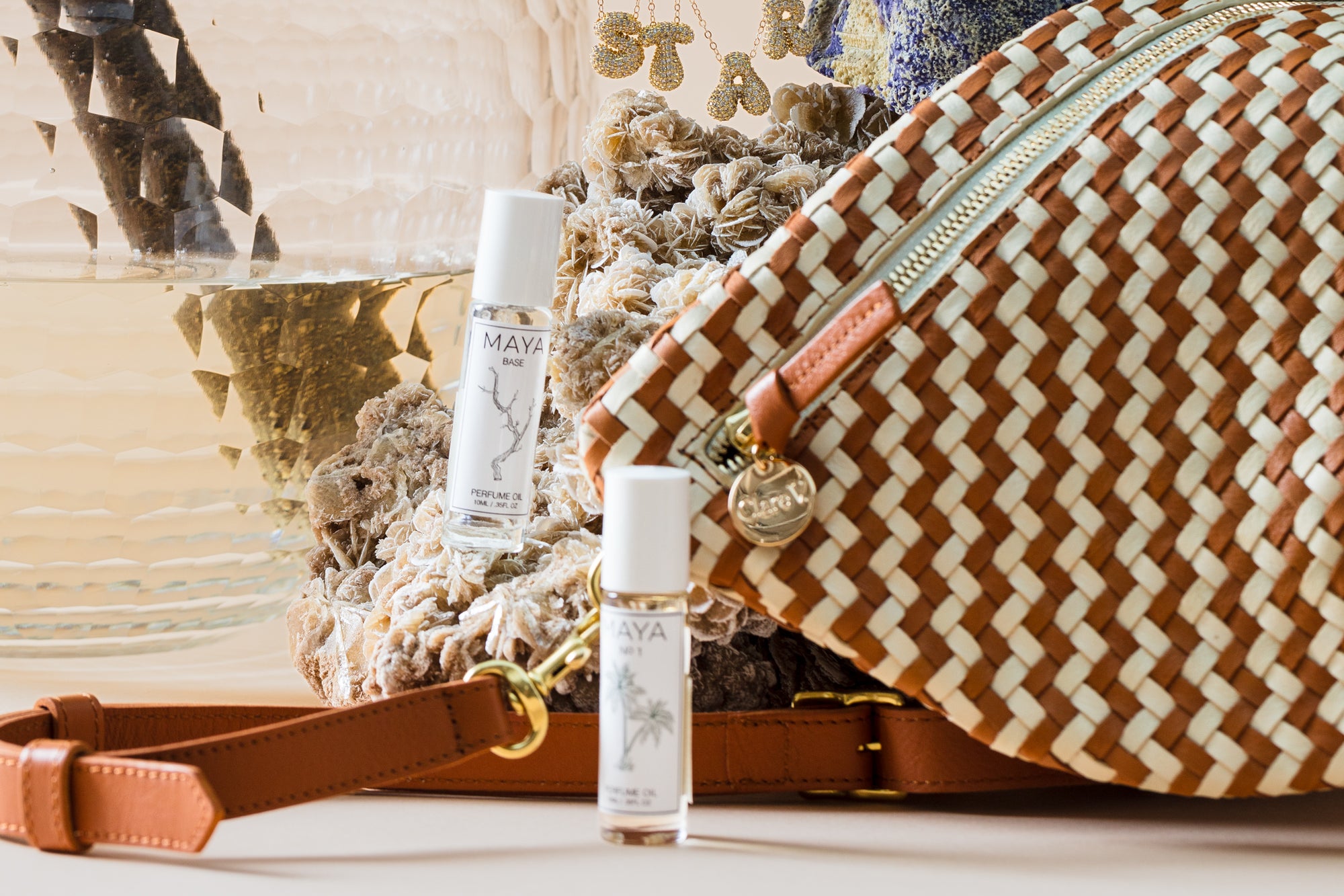 A woven bag with leather straps is placed next to two small bottles of "Maya" perfume. Behind them are dried flowers and a clear glass vase. A gold zipper and metal tag add detail to the bag.