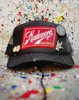 The MadeWorn Hat by Made Worn is a black trucker cap with a custom "Madeworn Art Dept" front patch, limited edition pins (dice, hand sign, and “The best things in life aren't things”), an adjustable back strap, and a paint-splattered backdrop.