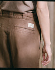 Someone wearing Hey Gang’s The Work Pant in brown cotton canvas—with a branded patch on the back pocket—stands by a wooden wall, their right arm relaxed at their side.