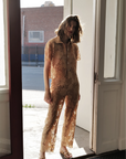 A woman stands in a doorway wearing Le Superbe Suntan Camp Pants and a sheer, floral lace shirt, sunlight streaming in behind her to create an effortlessly bohemian look.