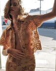 A woman with wavy, shoulder-length hair stands by a city street in sunlight, wearing the Le Superbe Suntan Camp Shirt—a nude floral lace blouse with a scalloped hem—styled partially unbuttoned and paired with high-waisted pants.