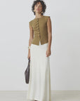 A person with short curly hair and earrings wears the SUNCOO PAGNI S/L button front brown sleeveless jumper and a long white skirt, holding a dark handbag and wearing bracelets, standing against a plain background.