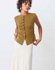 A woman models the SUNCOO PAGNI S/L button front jumper in olive green with an asymmetrical cut, paired with a long white skirt, against a plain white background, gazing slightly to the side.