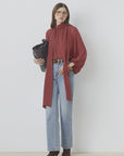 A woman stands against a white wall wearing the SUNCOO LINCOLN blouse with balloon sleeves and a bow collar, light blue high-waisted jeans, brown heeled boots, glasses, and holding a large black bag.