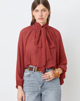 A woman poses against a white background wearing the SUNCOO LINCOLN blouse in rust muslin with a bow collar, paired with high-waisted light blue jeans and accessorized with hoop earrings, rings, and a chunky bracelet.