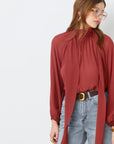 A woman wearing large glasses and the SUNCOO LINCOLN blouse—rust-red muslin with a scarf detail and balloon sleeves—paired with light blue high-waisted jeans and a brown belt, stands against a white background, looking down and to the side.