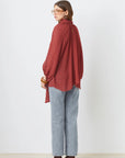 A woman stands facing away against a white background, wearing the SUNCOO LINCOLN blouse with balloon sleeves, light blue jeans, brown heels, and large glasses.