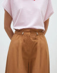 A person stands with their hands behind their back wearing brown cotton poplin Woven Pantalon Jayden by SUNCOO, paired with a pink and white striped short-sleeve top.