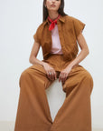 A woman sits on a stool wearing SUNCOO’s Woven Pantalon Jayden wide-leg pants and a brown short-sleeve button-up shirt, accessorized with a red scarf, pink-striped top, white studded shoes, and silver rings as she looks confidently at the camera.
