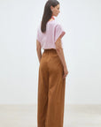 A woman stands indoors with her back to the camera, wearing SUNCOO's Woven Pantalon Jayden in brown organic cotton, paired with a pink and white striped short-sleeve top and cream shoes against a plain white wall.