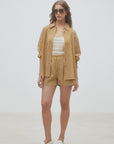A woman stands against a plain light background wearing SUNCOO's Woven Short Branden, styled with a yellow and white vertically striped shirt, white cropped top, sunglasses, and white loafers.