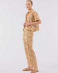 A person stands against a plain light background, wearing a tan floral lace short-sleeve shirt, Le Superbe Suntan Camp Pants, and tan sandals. With slicked-back hair and a neutral expression, they exude effortless bohemian romance.