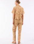 A person stands facing away, wearing Le Superbe Suntan Camp Pants with a tan short-sleeve lace top and flat sandals. Their hair is in a neat bun, radiating effortless bohemian style against a plain white background.