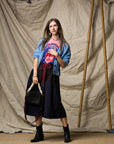 A woman poses before a canvas backdrop wearing a light blue cardigan, pink graphic tee, black ankle boots, a black braided-strap handbag, and the Le Vintage Sport Skirt by Ping Pong Surf Club.