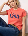 A young woman in the Clare Vivier Classic Tee CIAO—coral, short-sleeved, with bold blue lettering—pairs it with blue jeans and a tan belt. Made of lightweight cotton, she sits on the floor, smiling softly with her head resting on one hand.