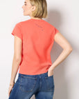 A woman with short blonde hair stands with her back to the camera, one hand in her pocket, wearing Clare Vivier's Classic Tee CIAO in coral and blue jeans against a plain white background.