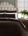A cozy bedroom features a decorative upholstered headboard, John Robshaw’s Velvet Coverlet King in dark brown with matching pillows, and a round wood bedside table topped with a glass vase of green foliage and stacked books by the window.