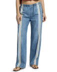 A person is seen from the waist down wearing Rag & Bone ftwt logan pull on track jeans in blue with vertical white side stripes, paired with a rust cropped top and beige sandals.