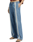 A person wears Rag & Bone ftwt logan pull on track jeans—relaxed straight fit, cotton-blend denim with two vertical white stripes, elastic waistband—paired with a rust crop top and sandals. Only their lower half is visible, hands in pockets.