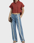 A woman wears a short-sleeve red cropped button-up shirt, Rag & Bone ftwt logan pull on track featherweight denim relaxed straight fit jeans with white side stripes, brown sandals, and holds a small dark clutch. One hand is in her pocket.