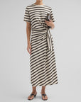 A person stands against a plain light background wearing the slub stripe tee midi dress by Rag & Bone, styled with a knotted waist, holding a small clutch bag and wearing black sandals.