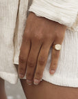 A close-up of a hand with water droplets, wearing a textured white garment and Faire's gold plated Everything Will Be OK Ring with engraved text. The background is softly blurred.