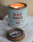 A lit G&W Soy Candle by Faire from the Happy Place Collection rests on an open book with a map beside a vintage compass, evoking nostalgic joy and inspiring thoughts of adventure and exploration.