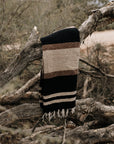 The TT Handwoven Blanket by Faire, featuring black, brown, and beige stripes, is draped over a weathered tree branch in a dry, brush-filled outdoor setting.