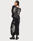A person with braided hair stands facing away, wearing the Me369 Beatrice Blouson Top with a black magic print and white tiger and sun designs, paired with a matching skirt and black chunky boots.