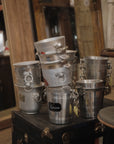 A stack of Go Home Vintage Champagne Buckets with handles sits on a dark wooden chest in a rustic room, surrounded by wooden furniture and mirrors.