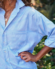 A person with curly hair wears the Frank & Eileen Eileen S25, a light blue patterned Italian cotton button-down, sleeves rolled up. With jewelry and hand on hip, the relaxed fit stands out against a blurred outdoor background.