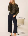 A woman stands indoors wearing the ASKK printed cropped long sleeve shirt, styled with black wide-leg pants and black slip-on shoes. She looks to the side with one hand near her mouth; a modern beige chair is behind her.