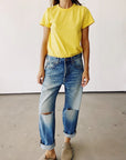At a vintage store, someone stands on a concrete floor wearing Hey Gang's yellow "The Crew" T-shirt made of 100% cotton and distressed blue jeans with rolled cuffs. Their hands hang by their sides above beige shoes, set against a plain light-colored wall.