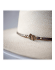 Close-up of the Lyndon Long Brim by Ninakuru, a beige woven hat made from sustainably harvested toquilla straw. It features a brown leather band and decorative knot detail, with its textured material and natural color highlighted against a softly blurred background.