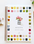 Faire's Watercolor Workbook is a beginner-friendly, spiral-bound guide featuring a flower vase illustration with colorful paint swatches around the border. Paint tubes, a brush, and a small plant accent the white surface nearby.