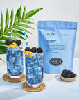 Two glasses of Faire Blackberry Jasmine Iced Tea sit on woven coasters. Next to them are a blue bag of tea sachets and a small bowl of blackberries, with a green leaf visible in the background.