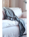 The Indaba Houndstooth Throw in teal, made from Indian cotton with fringed edges, is draped over a white couch in a cozy, softly lit living room, adding a stylish textured accent to the space.