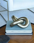 The HomArt Infinity Knot / Brass, a striking metallic decor piece, rests on a stack of books on a wooden table with a white chair featuring thin gold legs in the background.