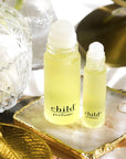 Two bottles of Faire Child Perfume Oil Roll sit on a marble tray, with a crystal jar and gold leaf accessory nearby, all bathed in bright natural light.