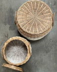 Two Faire Lidded Round Baskets with straw side handles rest on a gray concrete surface; one is closed, and the other reveals its woven white and natural interior. Both baskets combine style and function for versatile storage.