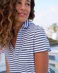 A woman with curly hair smiles and looks to the side while wearing the Frank & Eileen Charlotte Perfect Polo in navy and white stripes. She is outdoors, with a blurred background of buildings and sky.
