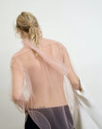 A person with long, blonde hair stands facing a light-colored wall, wearing the sheer pink Dear Polly Bow Blouse by Aquarius Cocktail. They hold a white garment over one arm and have a tattoo on their lower back.