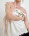 In a neutral setting, an individual is wearing the Aquarius Cocktail "Dear Polly Bow Blouse," a sheer peach-colored mesh tulle top with an oversized fit over a white garment with a pin, highlighting its delicate texture and casual style.