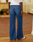 A person stands indoors on a tiled floor wearing Frank & Eileen’s NYC Trouser jean in blue denim, paired with a white long-sleeve blouse and white sandals. The photo is taken from behind, showing part of a kitchen in the background.