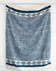 The Selene Momo Blanket by Jitana Wholesale is a blue and white woven textile, handcrafted by Mayan artisans. It has horizontal stripes, decorative floral bands near the edges, and slightly frayed ends.